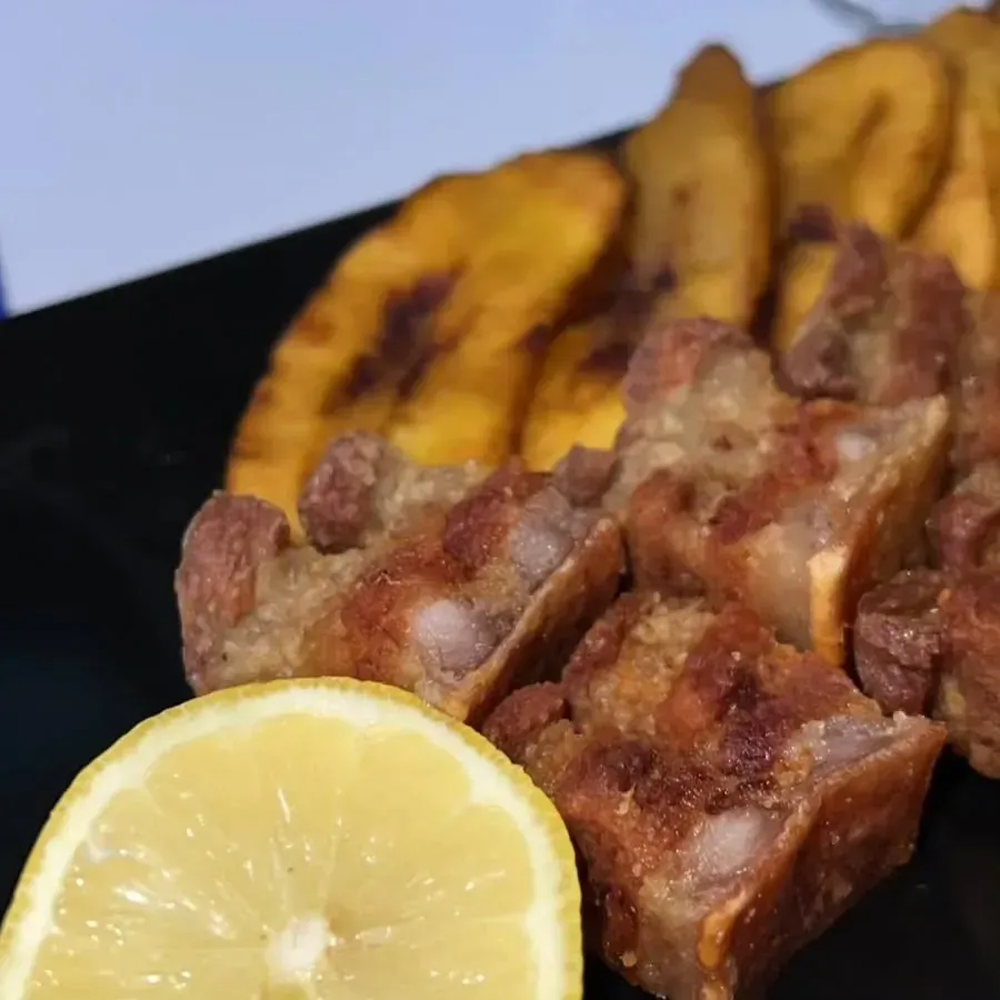 Panceta de cerdo frita crujiente con plátanos fritos y una rodaja de limón en un plato negro.