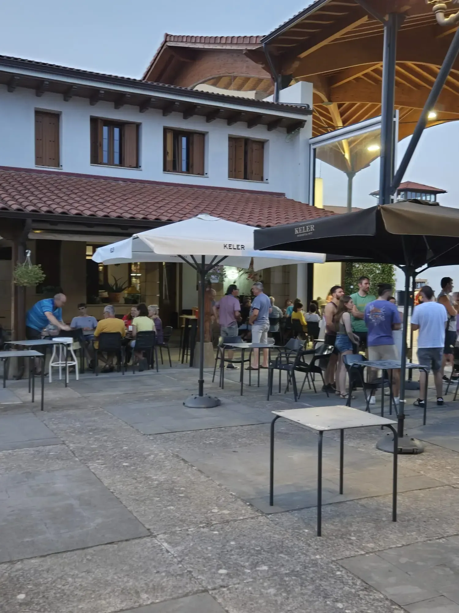Patio al aire libre con gente sentada en mesas, bajo sombrillas, cerca de un edificio blanco con techo marrón.