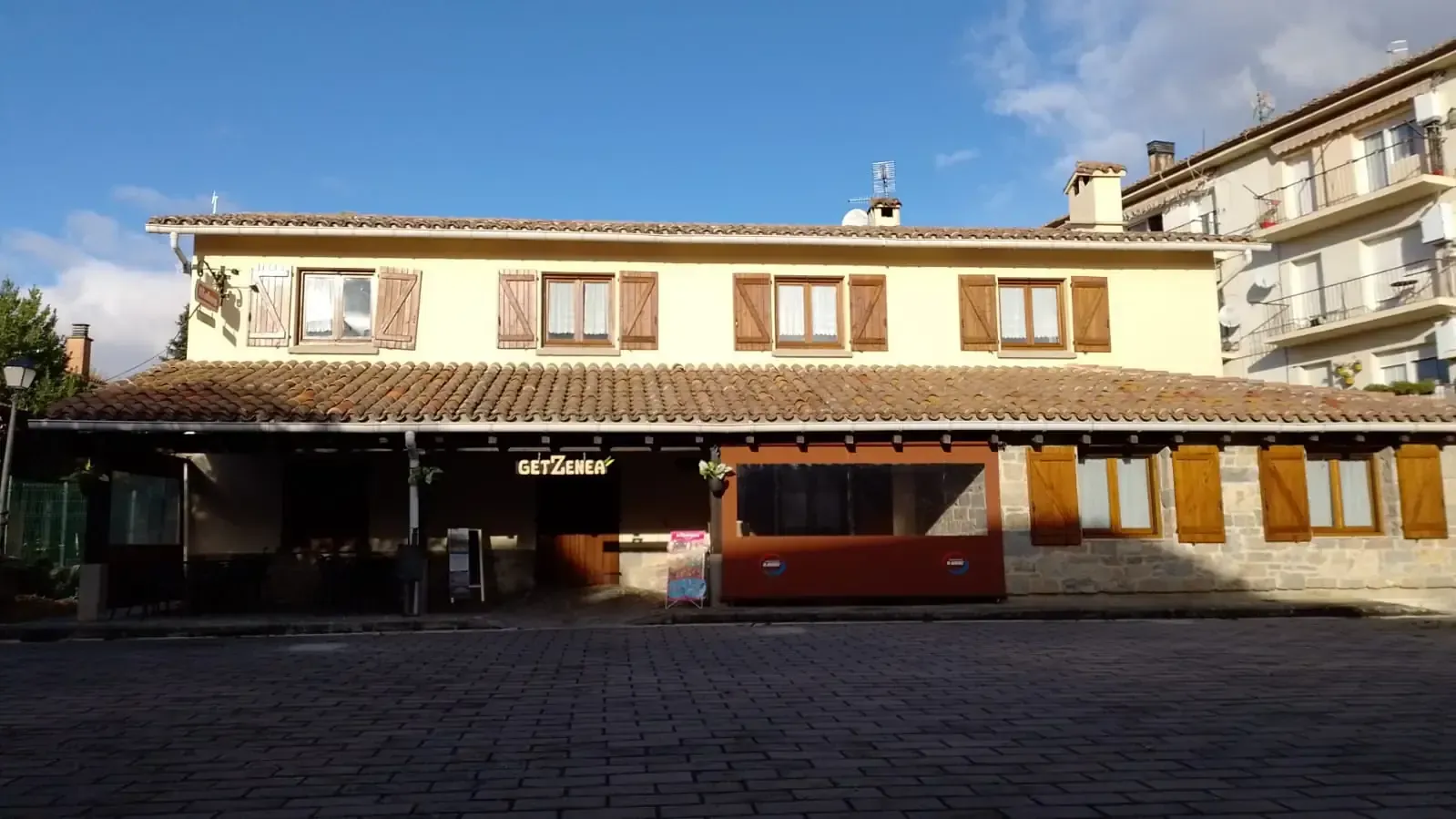 Edificio de dos plantas con fachada amarilla, persianas marrones y techo de tejas.