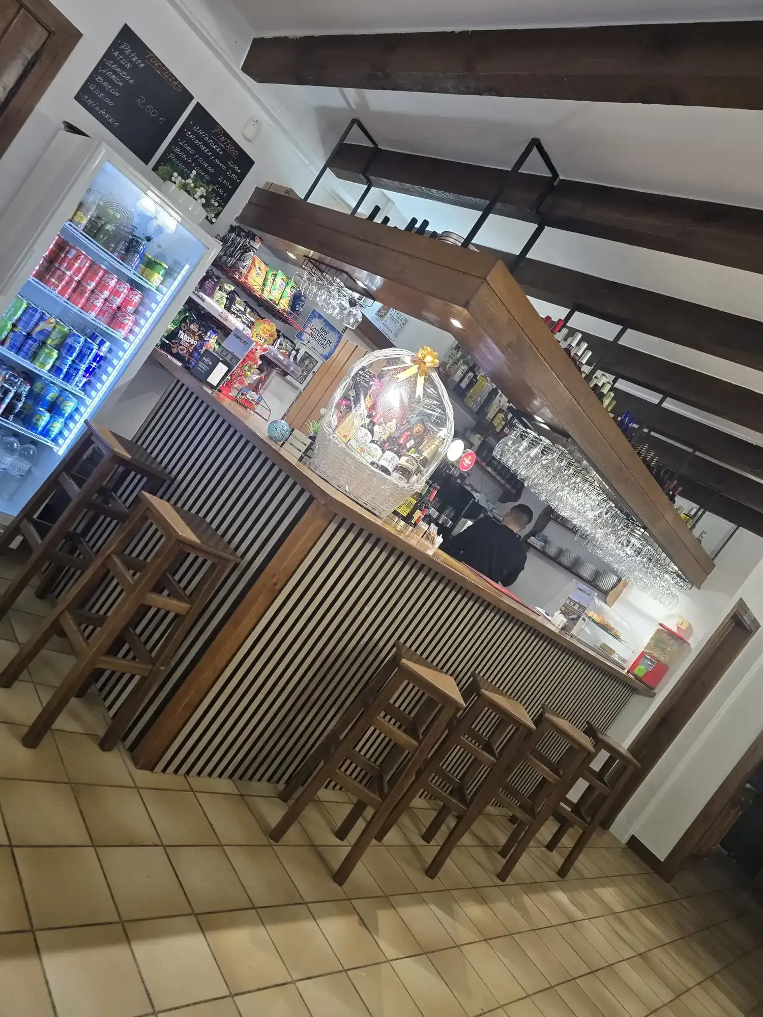 Barra con taburetes, frigorífico para bebidas y estantes con botellas; vigas de madera en el techo.