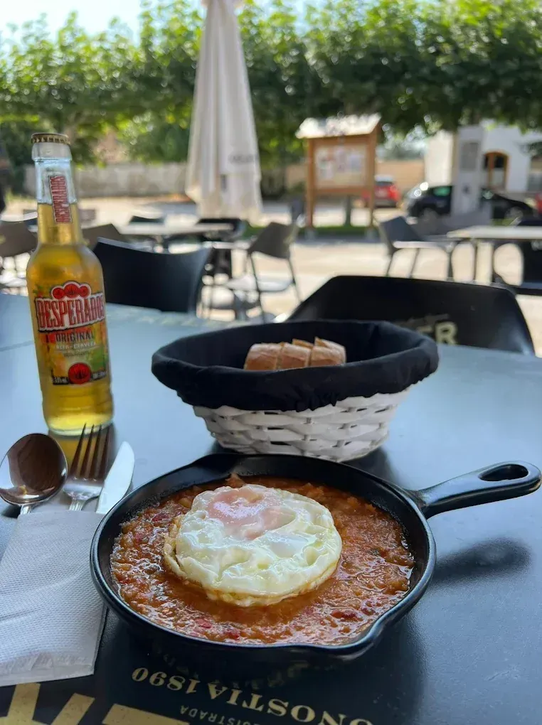 Un huevo frito con salsa en una sartén, pan y una cerveza en una mesa al aire libre.