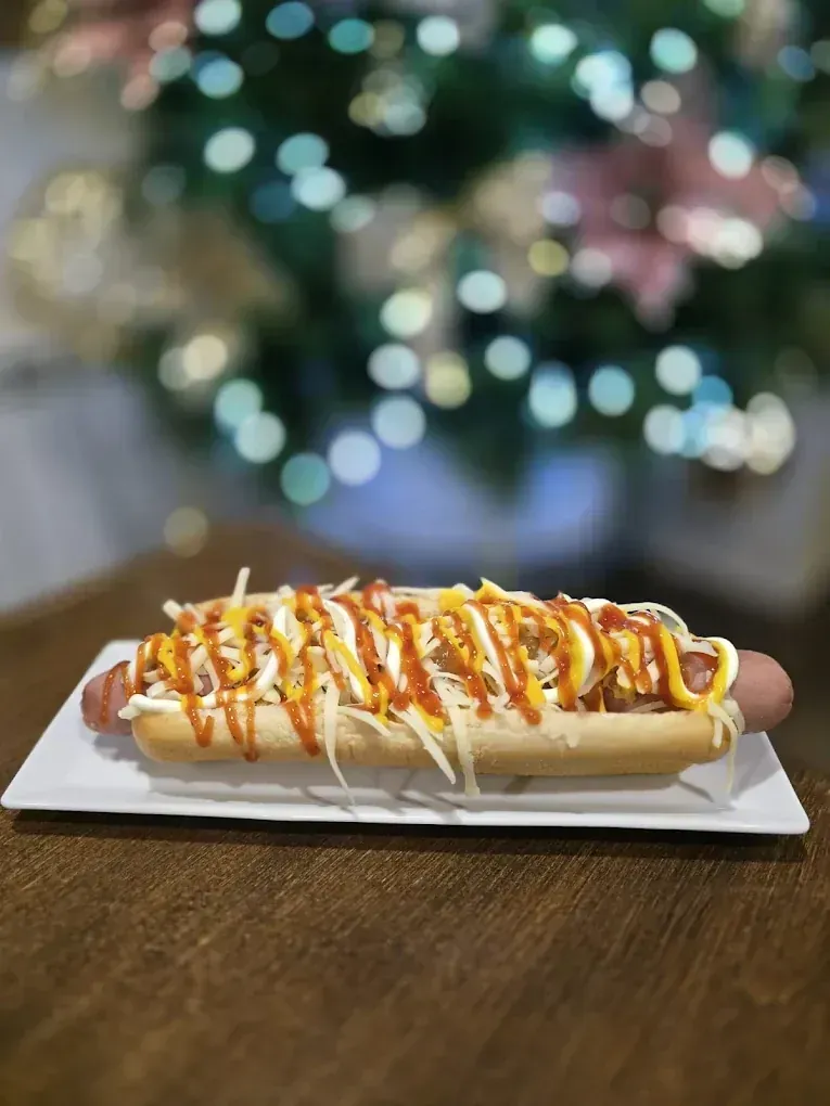 Perro caliente en un plato blanco, cubierto con queso rallado, mostaza y ketchup, fondo bokeh.