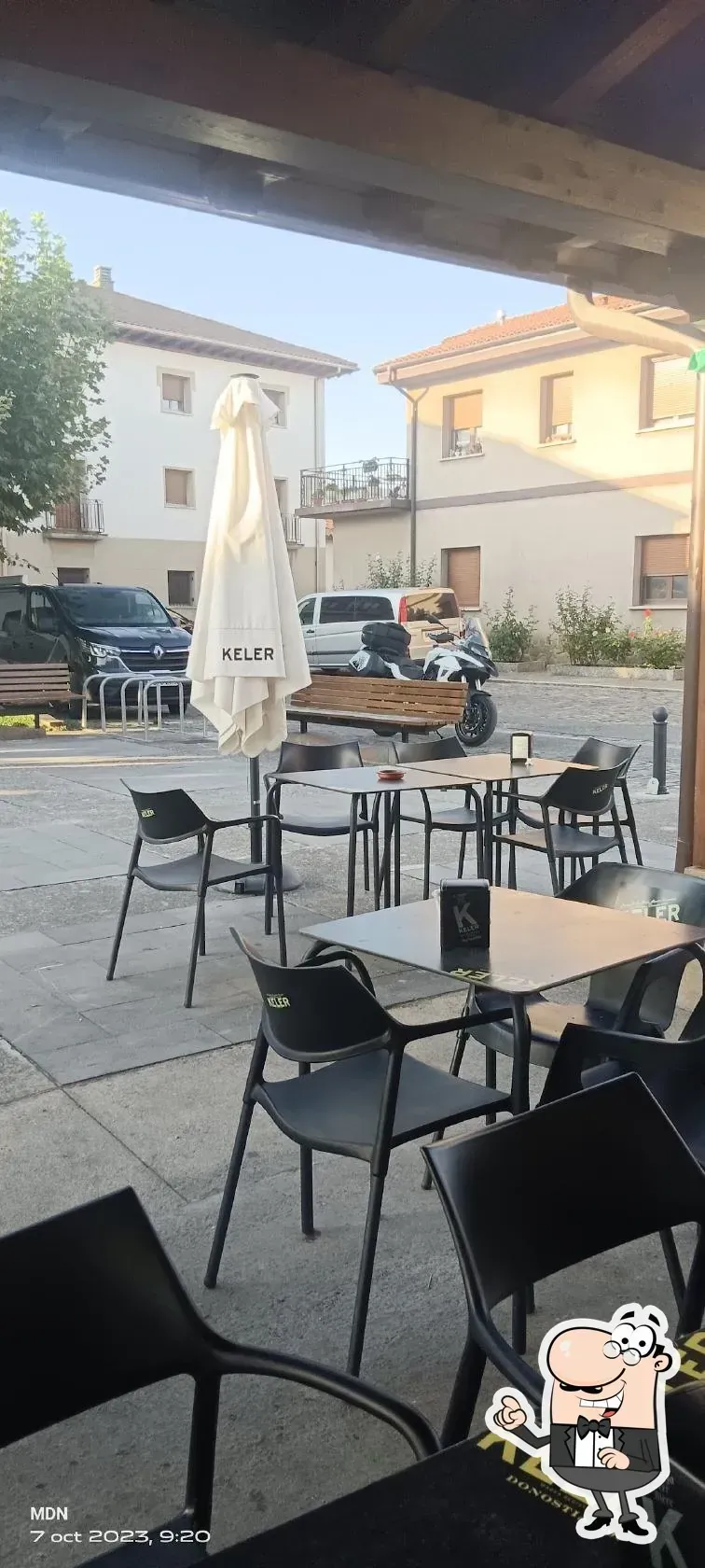 Cafetería al aire libre con mesas y sillas bajo un toldo de madera. Edificios y vehículos al fondo.