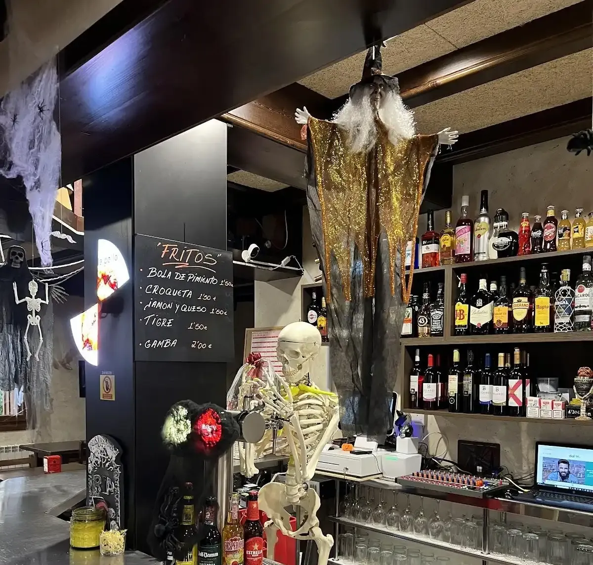 Bar decorado para Halloween con brujas colgantes, esqueletos, botellas de licor y un cartel iluminado.