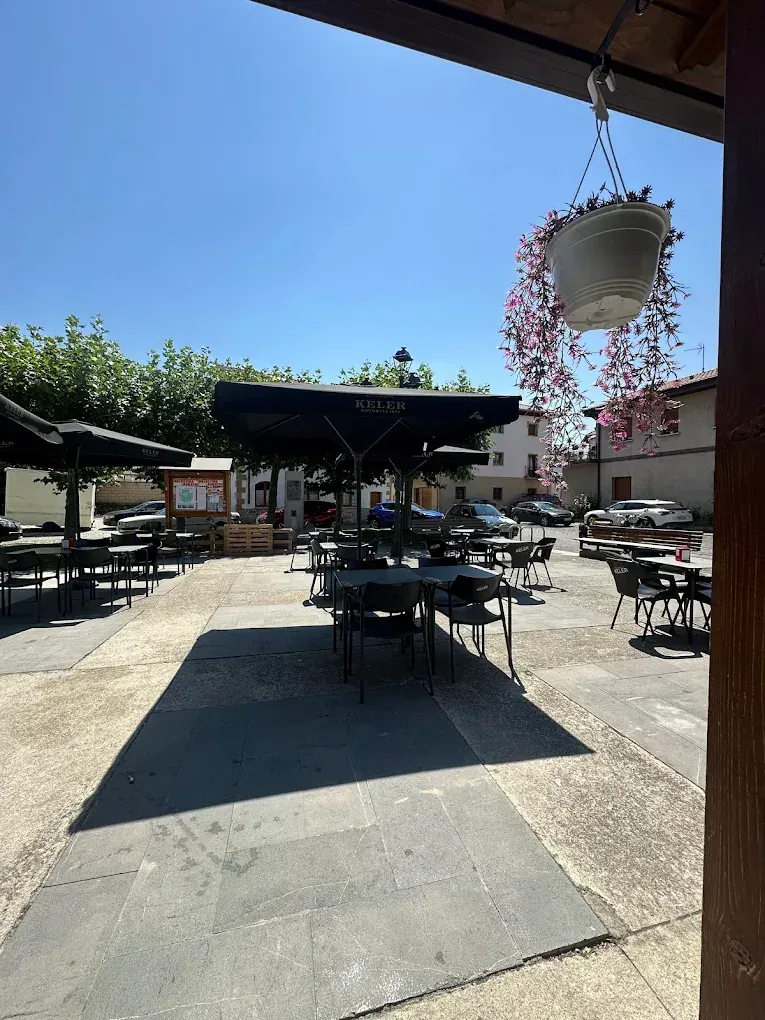 Mesas y sillas de café al aire libre en una plaza pavimentada con una canasta de flores colgante en un día soleado.