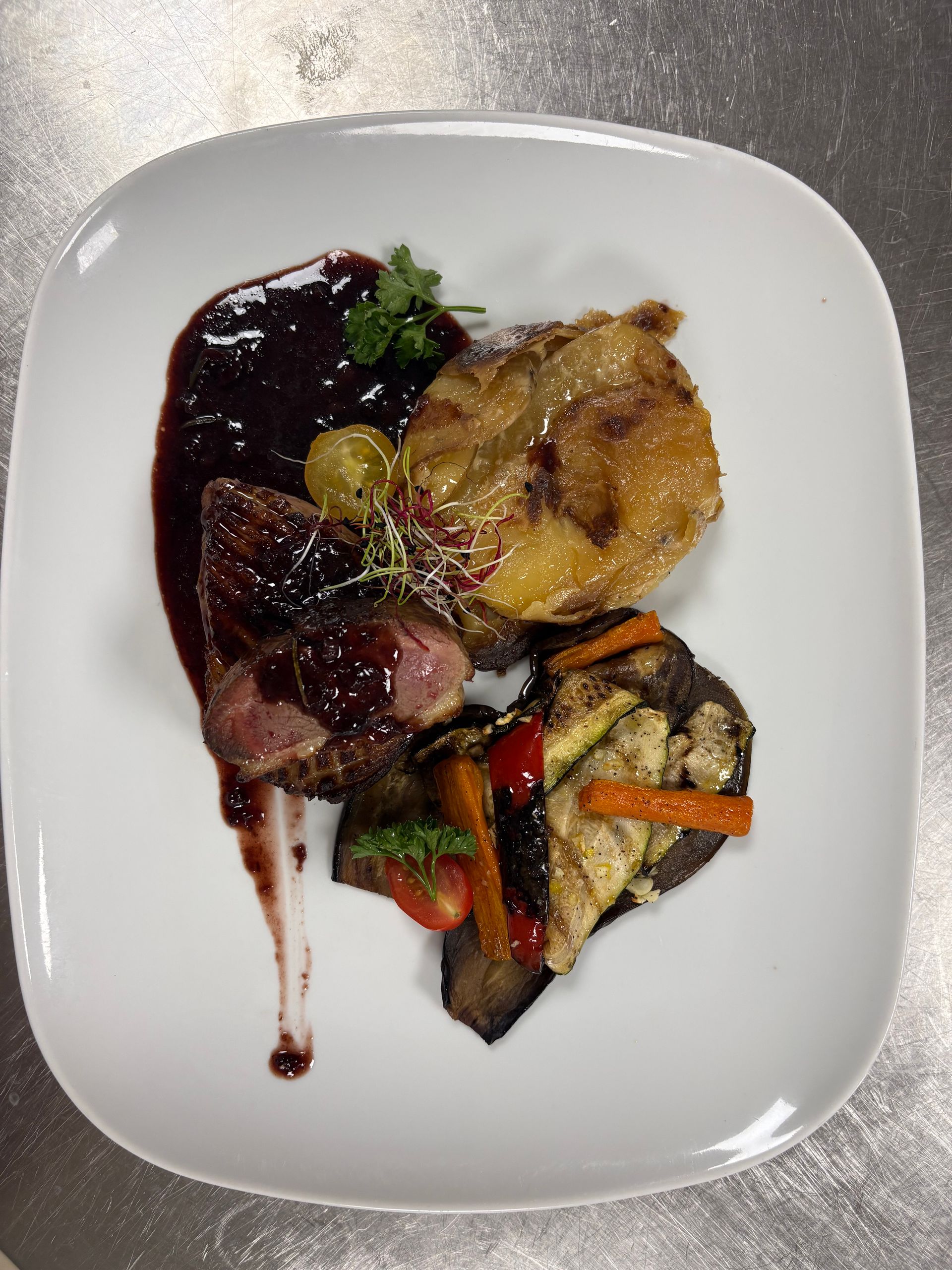 Plat gastronomique servi à l'assiette, avec sauce brune, pommes de terre rôties et légumes, dans une assiette carrée blanche.