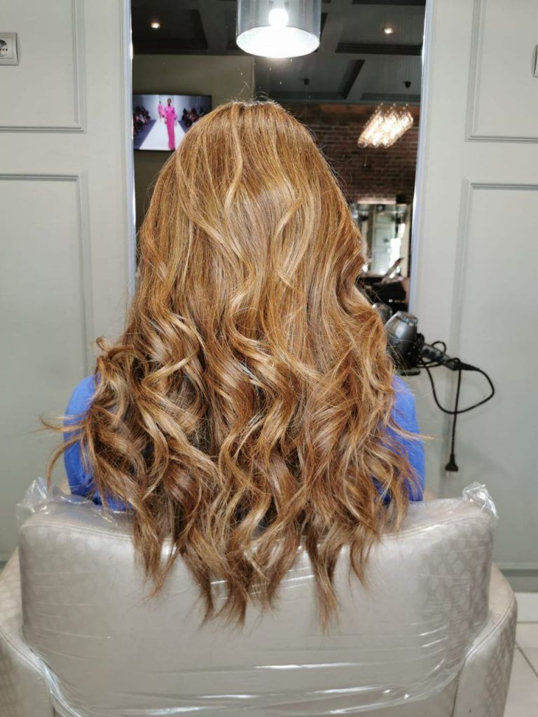 Mujer con cabello rubio largo y ondulado sentada en una silla de peluquería, vista desde atrás.