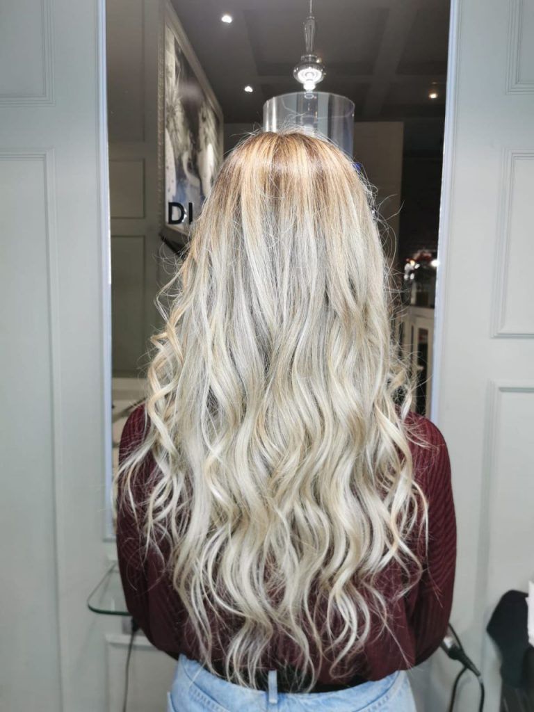 Persona sentada en una silla de peluquería con cabello rubio largo y ondulado, vista desde atrás en un puesto de peinado iluminado por un espejo.