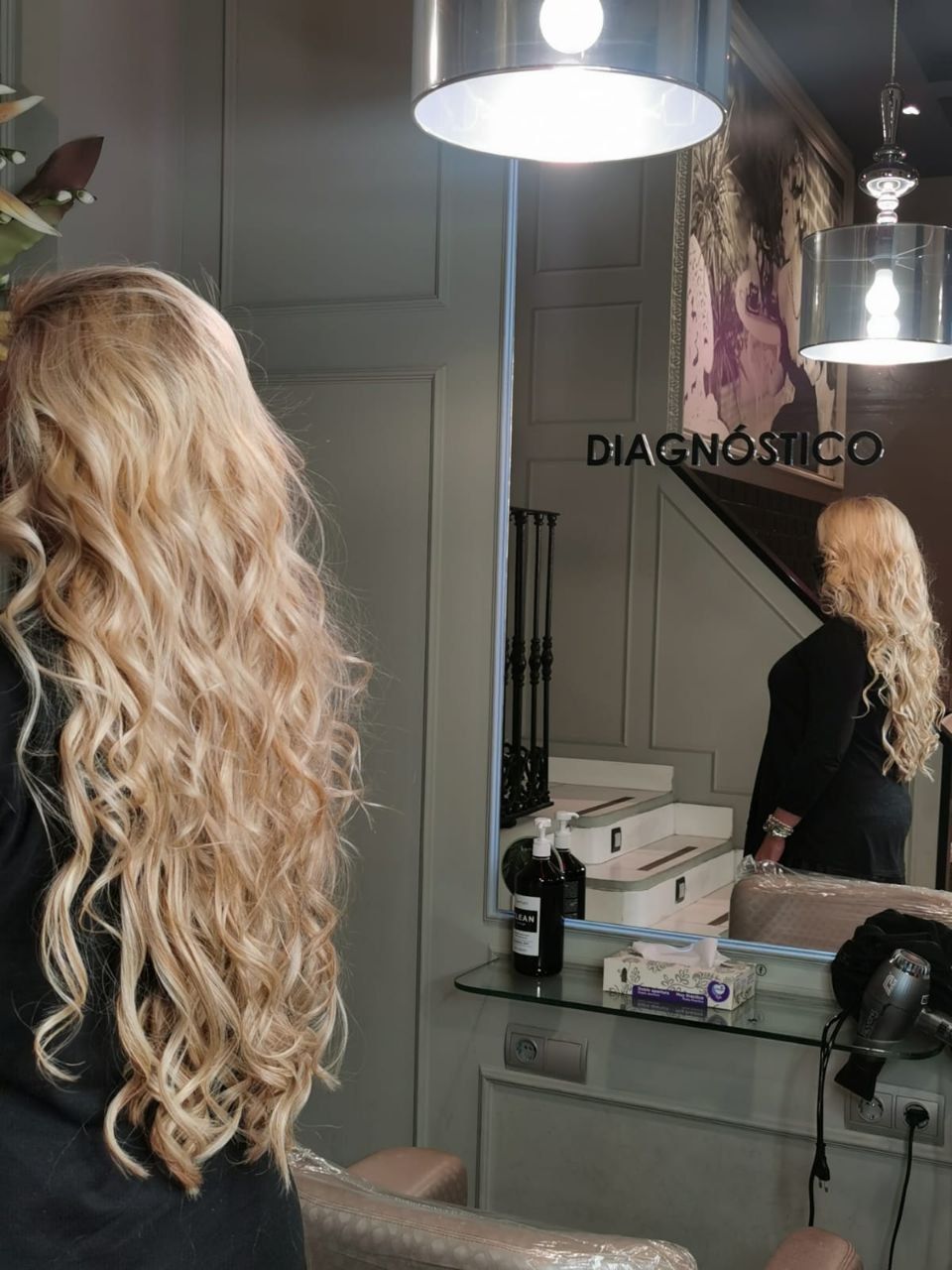 Mujer con cabello rubio largo y ondulado de pie en una peluquería, vista de espaldas junto a un espejo y un lavabo.
