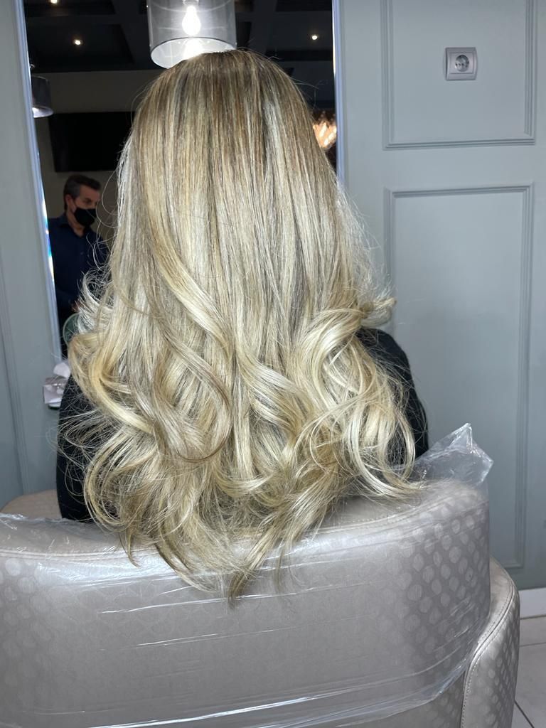 Persona con cabello rubio largo y ondulado sentada en una silla de peluquería cubierta con plástico, vista desde atrás.