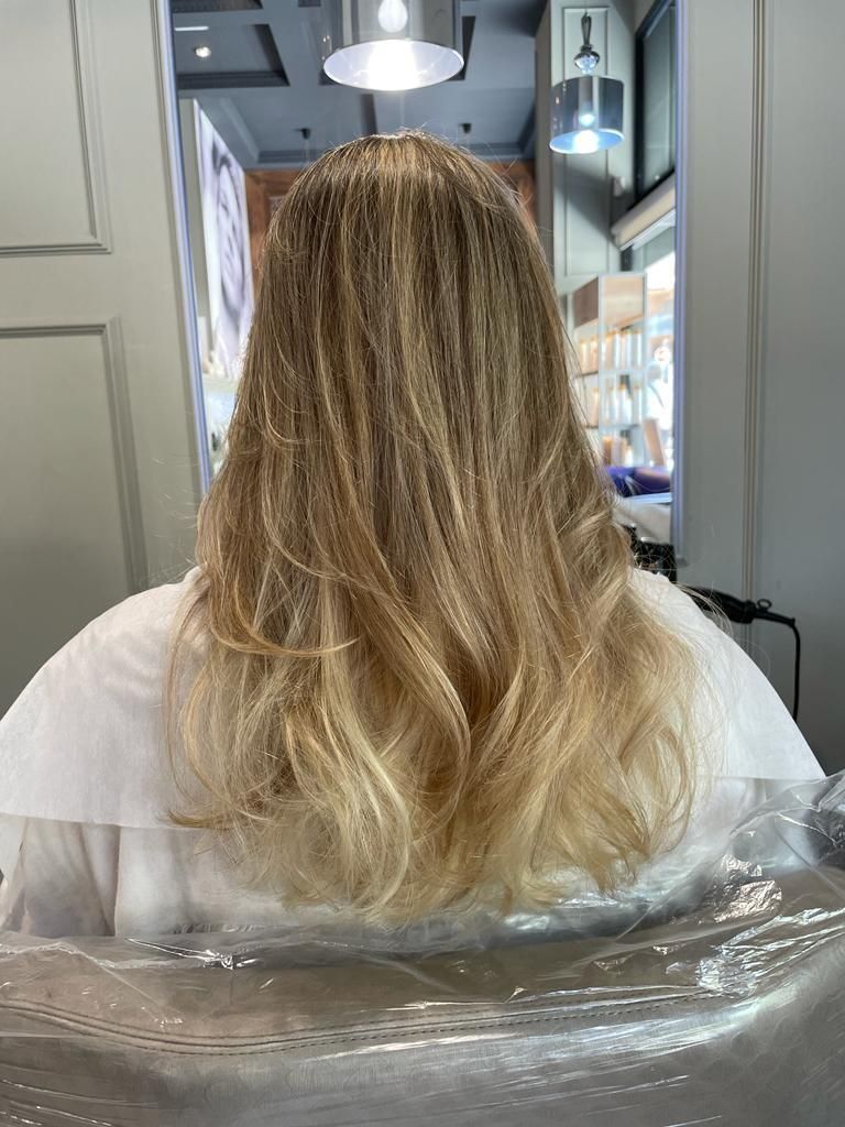 Vista trasera de una persona con cabello rubio largo sentada en una silla de peluquería, envuelta en una capa de plástico transparente.