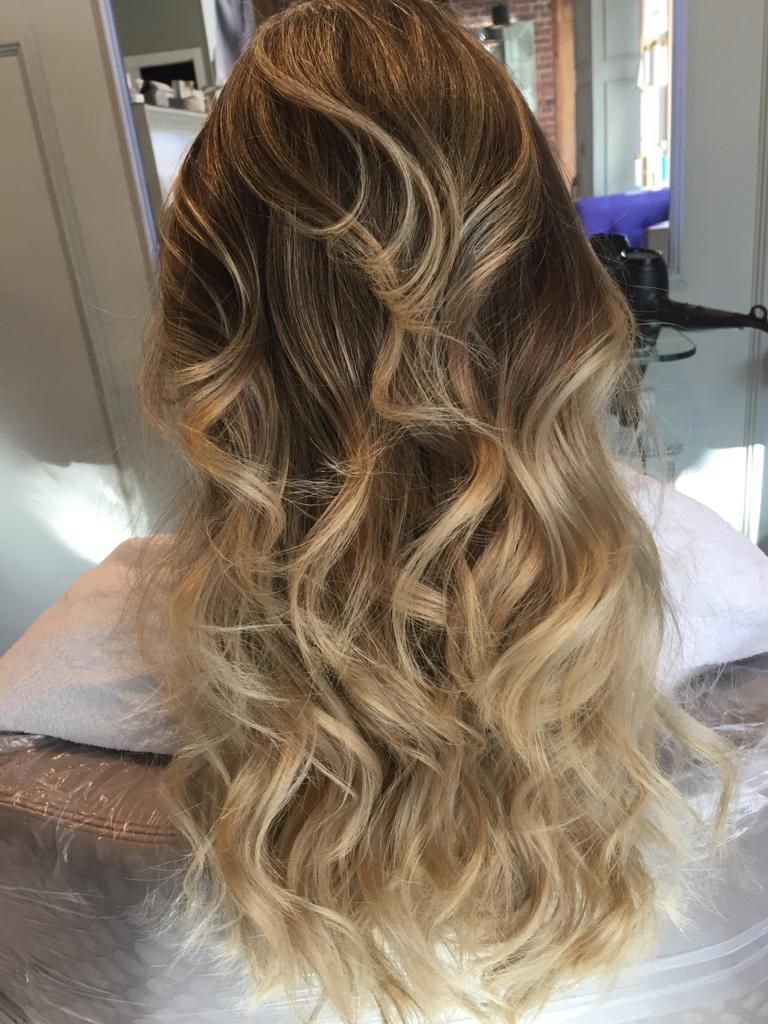 Mujer con cabello rubio largo sentada en una silla de peluquería, vestida con una capa blanca y cubierta con plástico transparente.