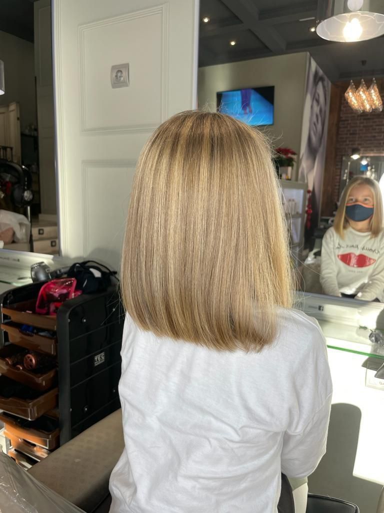 Una persona con cabello rubio hasta los hombros está sentada en una silla de peluquería frente a un espejo.