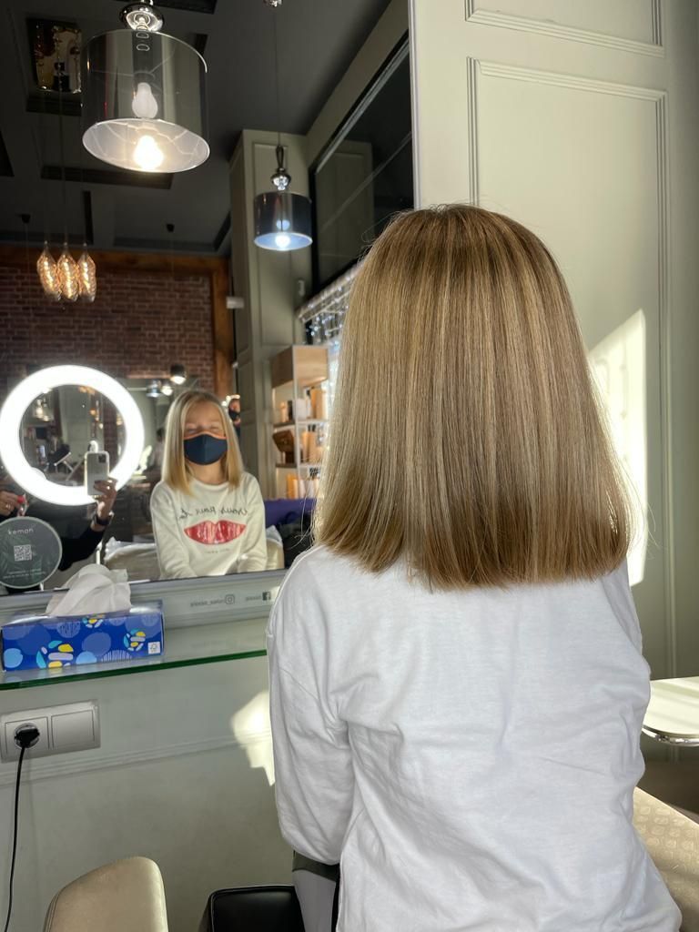 Una persona con cabello rubio hasta los hombros está sentada en una silla de peluquería frente a un espejo.
