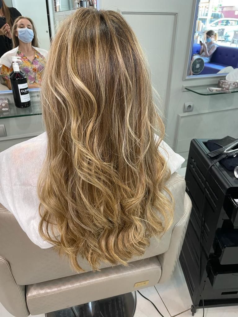 Mujer con cabello rubio largo y ondulado sentada en una silla de peluquería, vista desde atrás en un salón lleno de espejos.