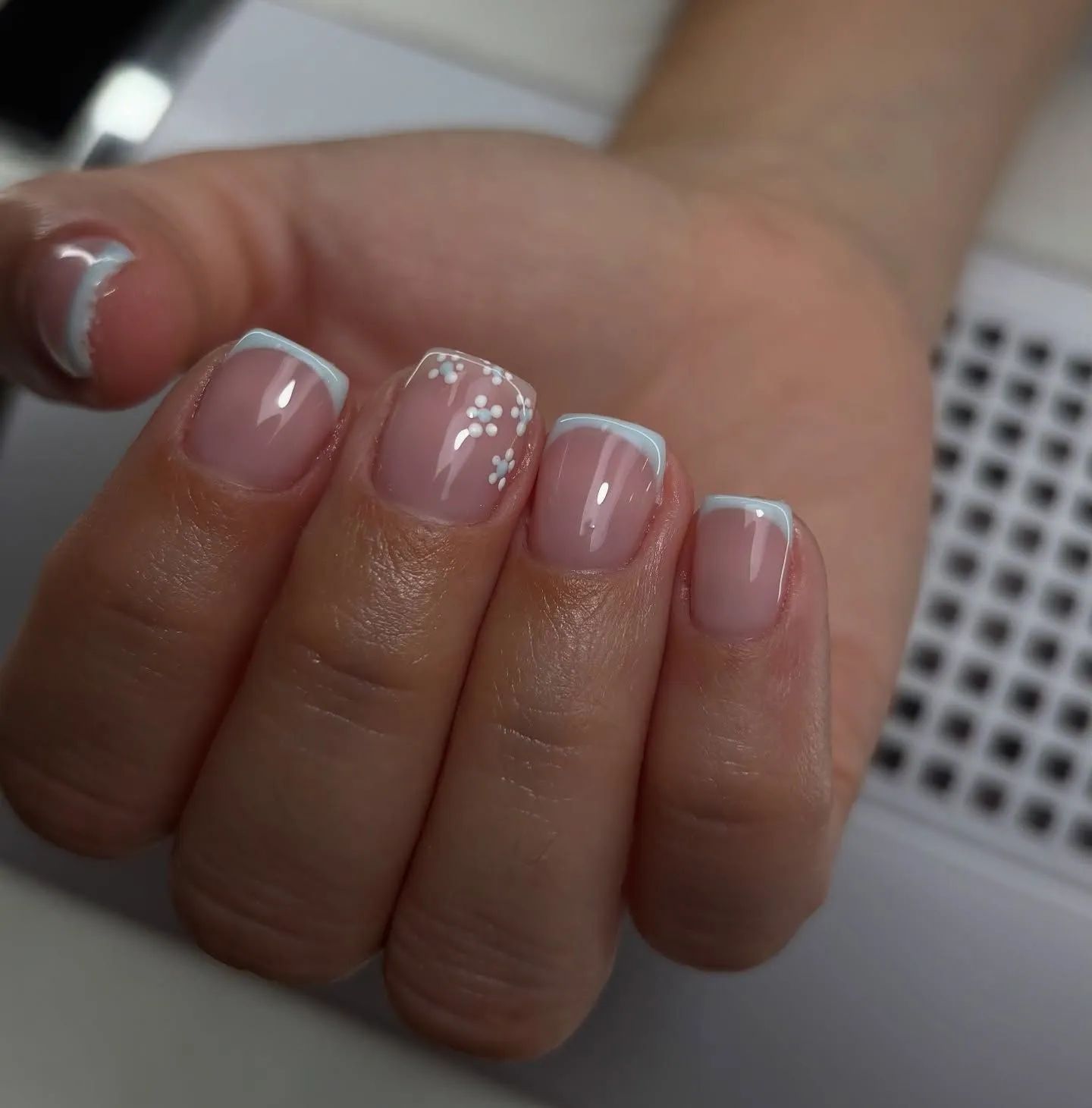 Mano con uñas de punta francesa, una de ellas decorada con pequeñas flores blancas.