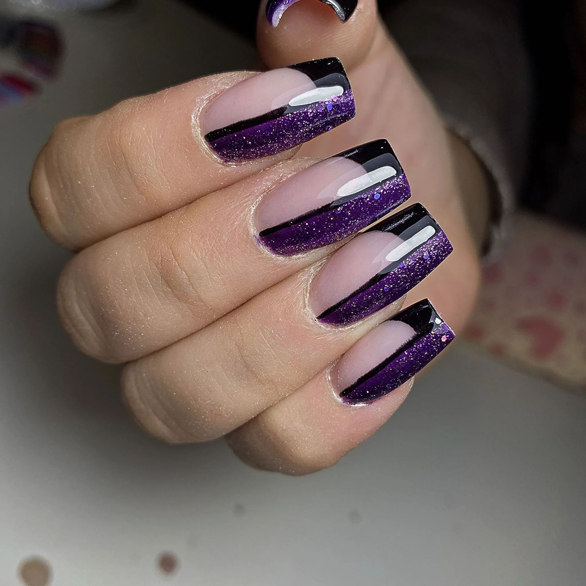 Mano con uñas de forma cuadrada; diseño de brillo negro y morado con punta francesa y raya diagonal.