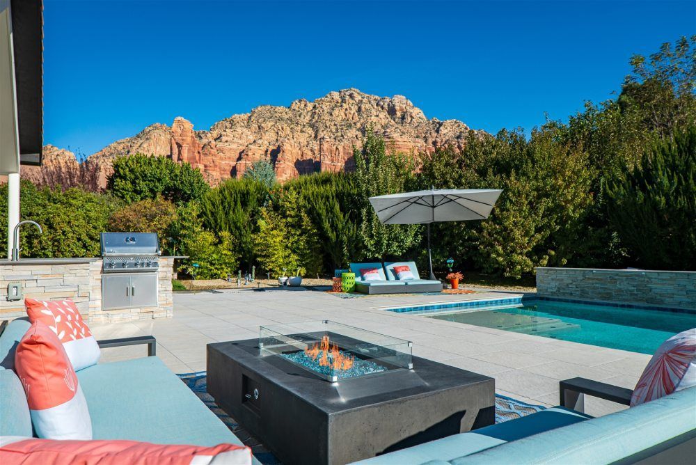 Outdoor patio with pool, fire pit, grill, and lounge furniture, set against red rock mountain backdrop under blue sky.