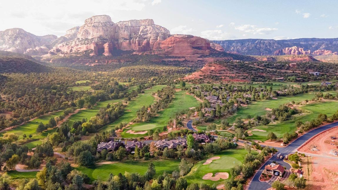 Golf course nestled in a valley with red rock mountains in the background, green fairways and trees.