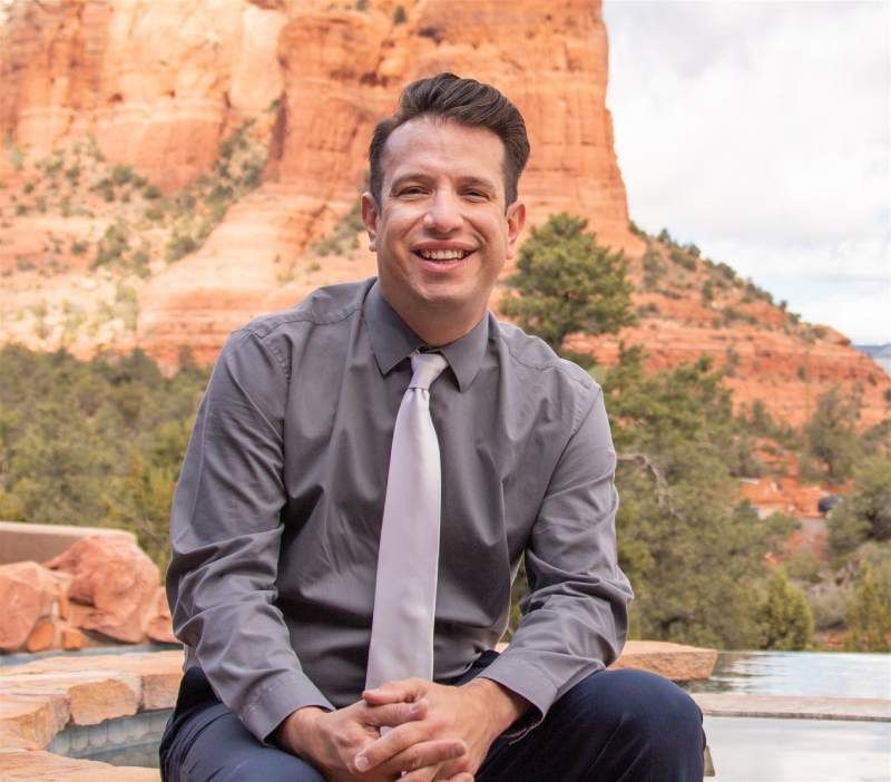 Man in a dark blue polo shirt smiles, posing outdoors with a mountain backdrop.