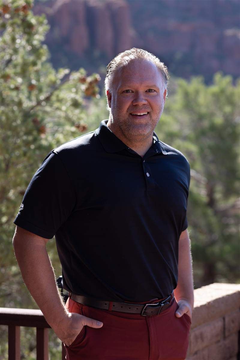 Man in a dark blue polo shirt smiles, posing outdoors with a mountain backdrop.