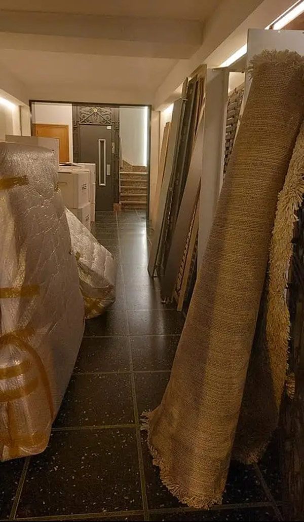 Un estrecho pasillo de almacenamiento revestido con alfombras enrolladas y muebles envueltos apoyados contra las paredes.