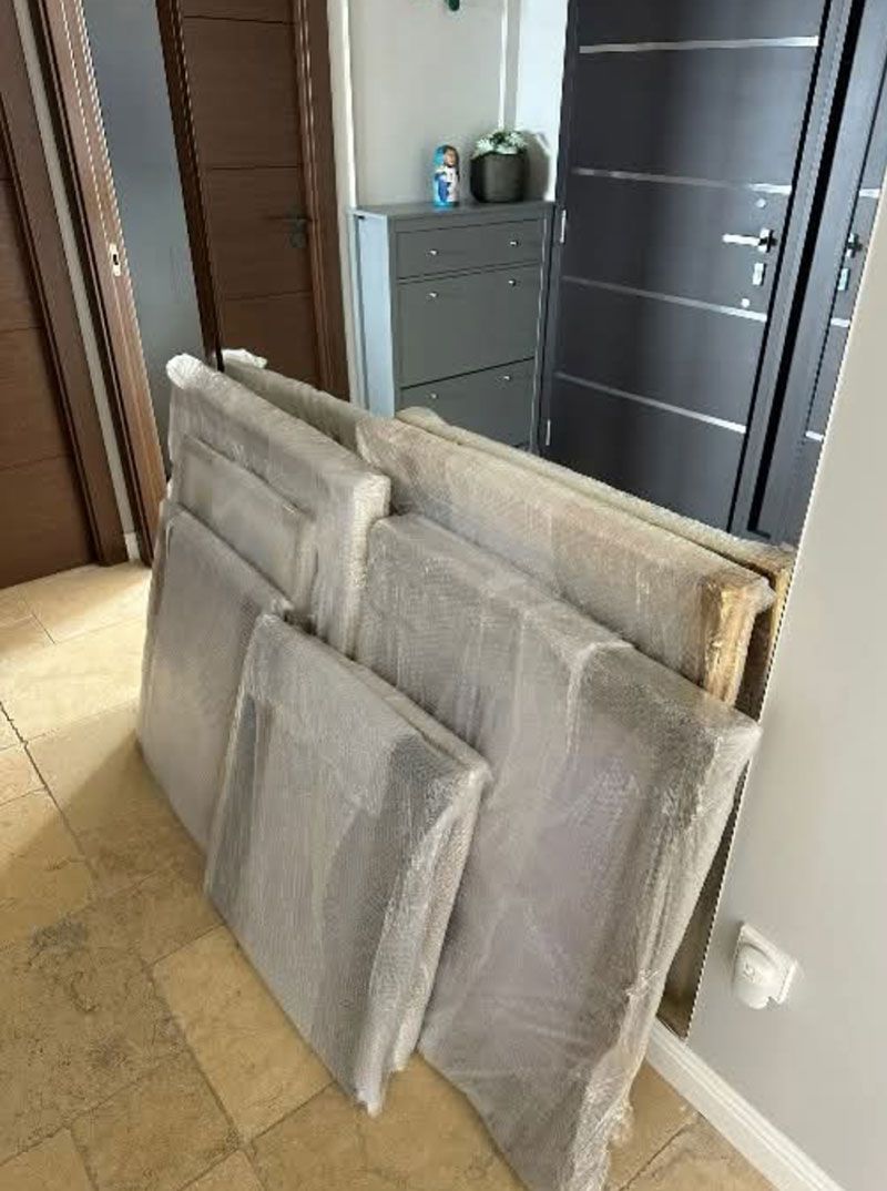Pila de paneles enmarcados apoyados contra una pared en un pasillo cerca de una cómoda y una puerta.