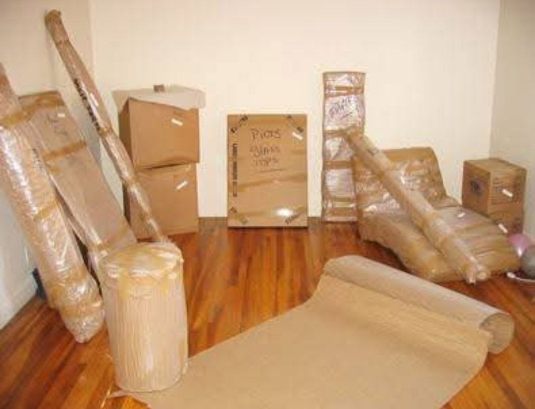 Cajas de cartón apiladas y muebles envueltos en una habitación con suelos de madera, como si se estuvieran mudando o guardando en un almacén.