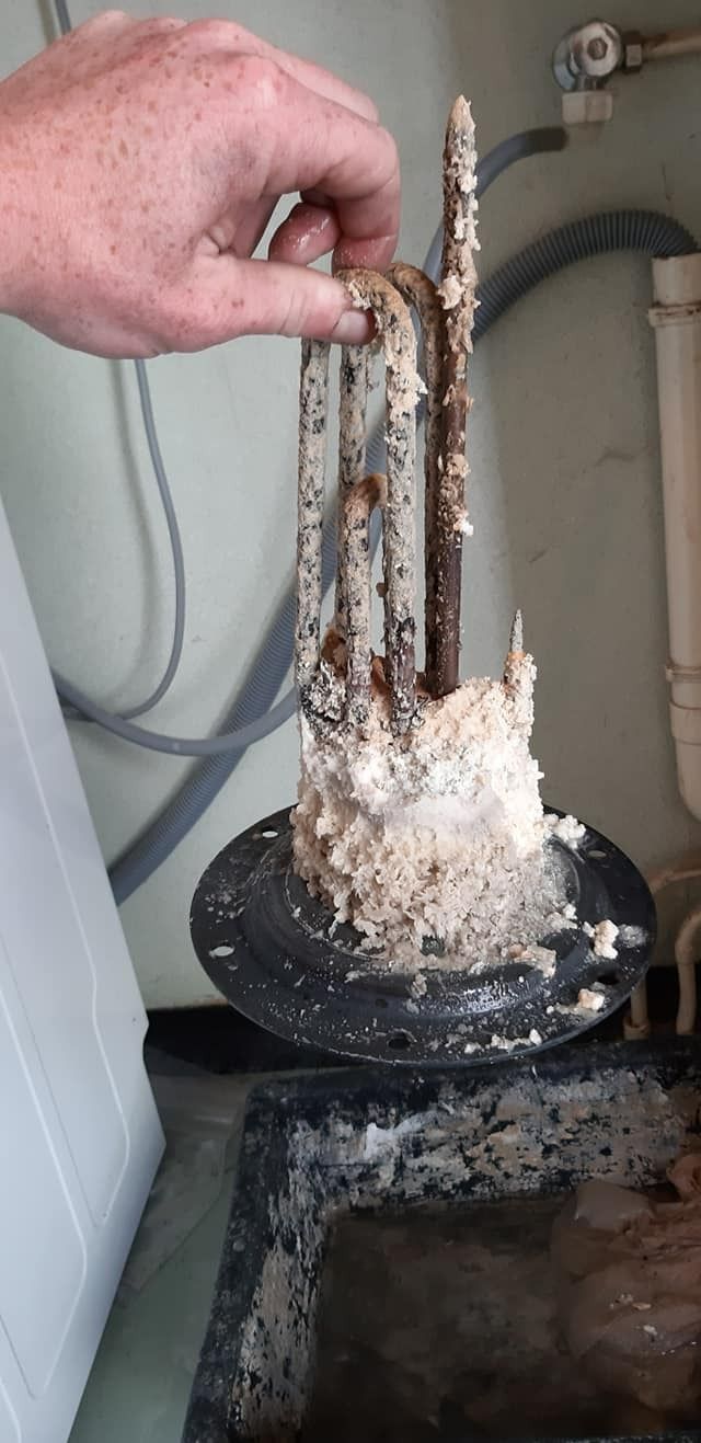 Une main tient un élément chauffant de chauffe-eau fortement incrusté de dépôts minéraux blancs, retiré d'un réservoir d'eau.