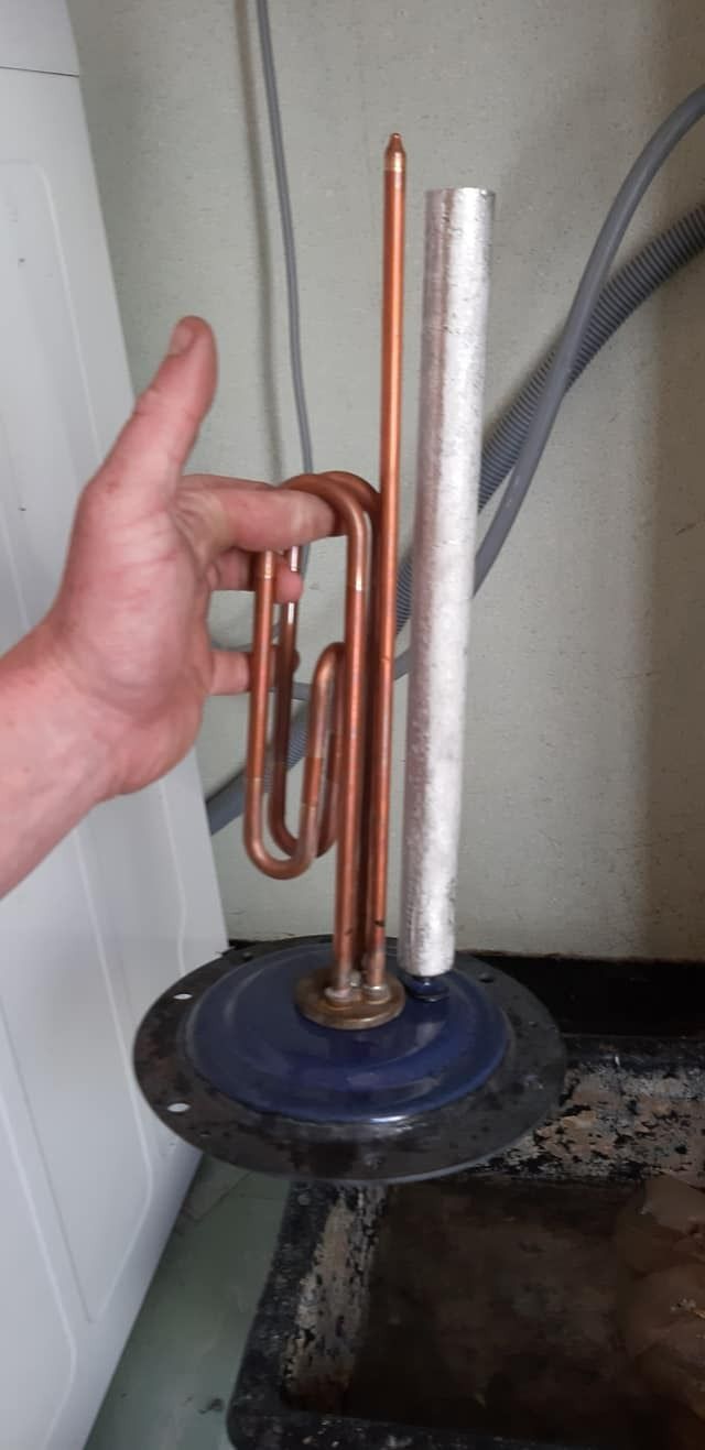 Une main tenant un élément chauffant de chauffe-eau et une tige d'anode montée sur une base métallique circulaire.