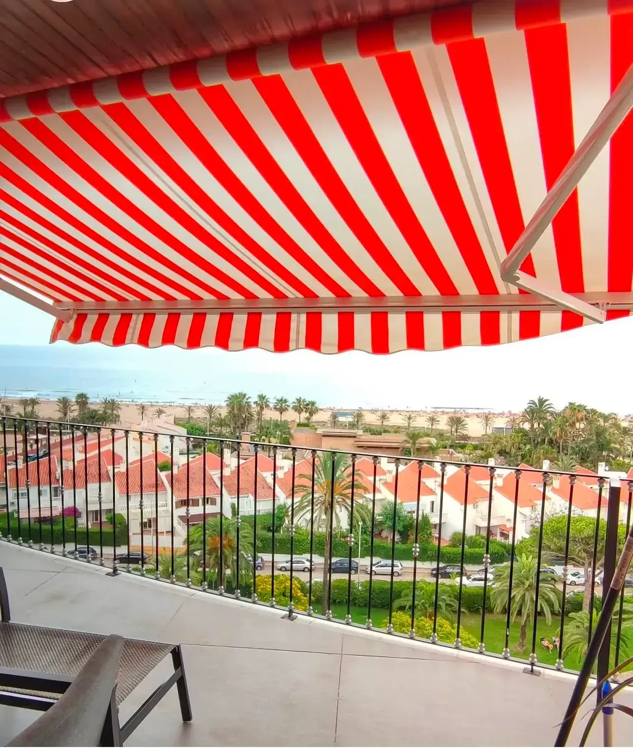Balcón con toldo de rayas rojas y blancas con vistas a un barrio residencial y zonas verdes.