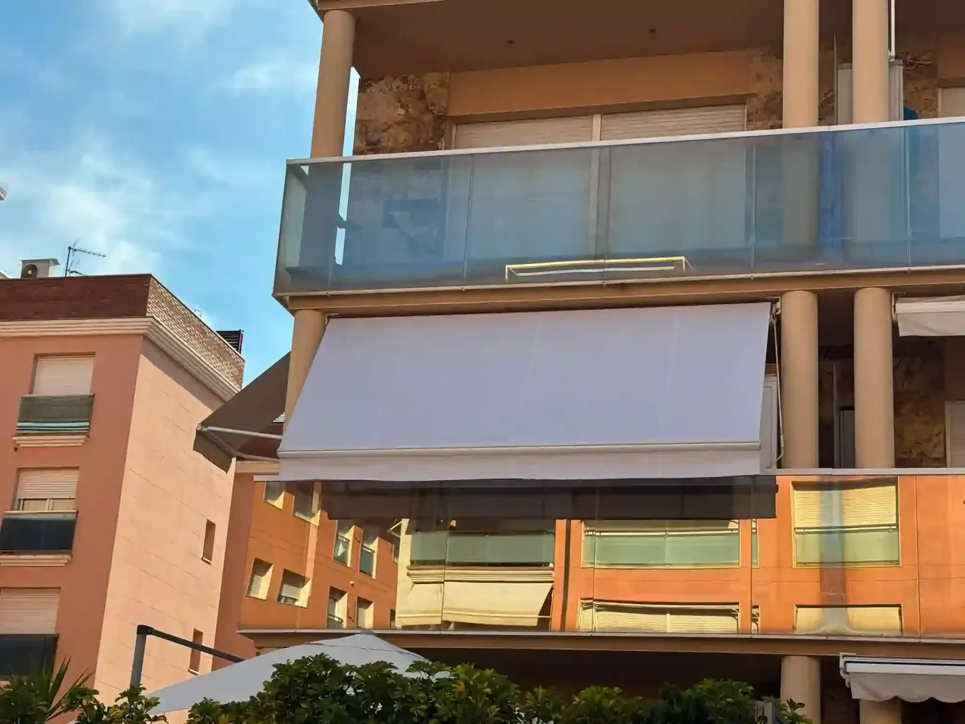 Edificio moderno con balcones de cristal y un gran toldo contra un cielo azul.
