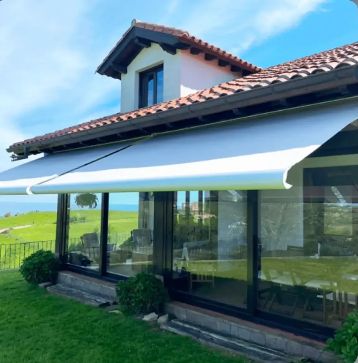Casa con toldo blanco sobre patio acristalado, tejado de tejas rojas y campos verdes al fondo.