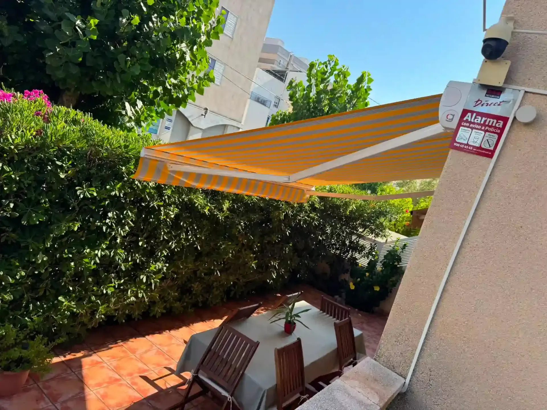 Patio con mesa bajo un toldo de rayas naranjas junto a setos y una pared beige.