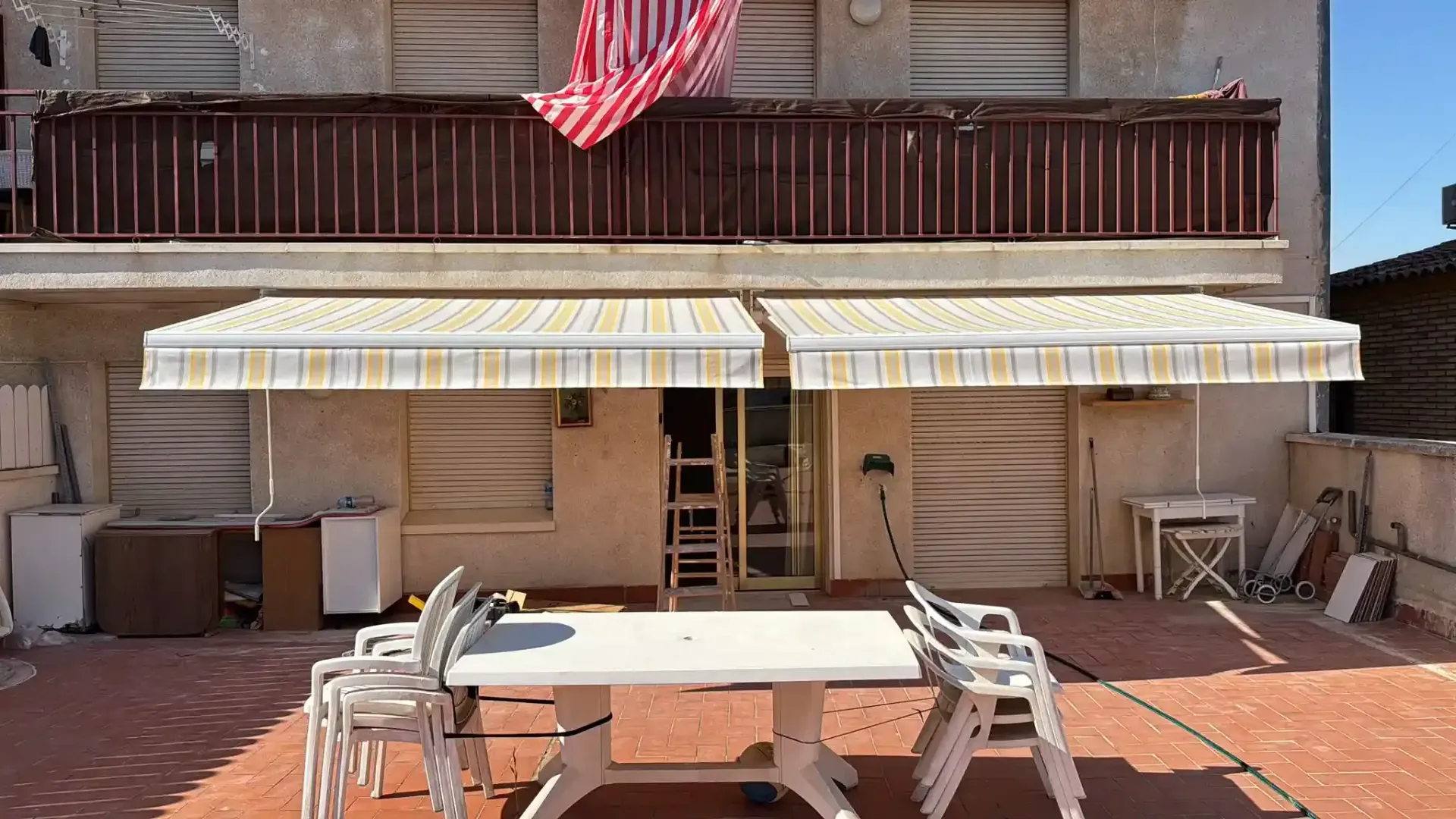 Patio trasero con mesa y sillas, toldos a rayas y una bandera estadounidense en una casa de dos pisos.