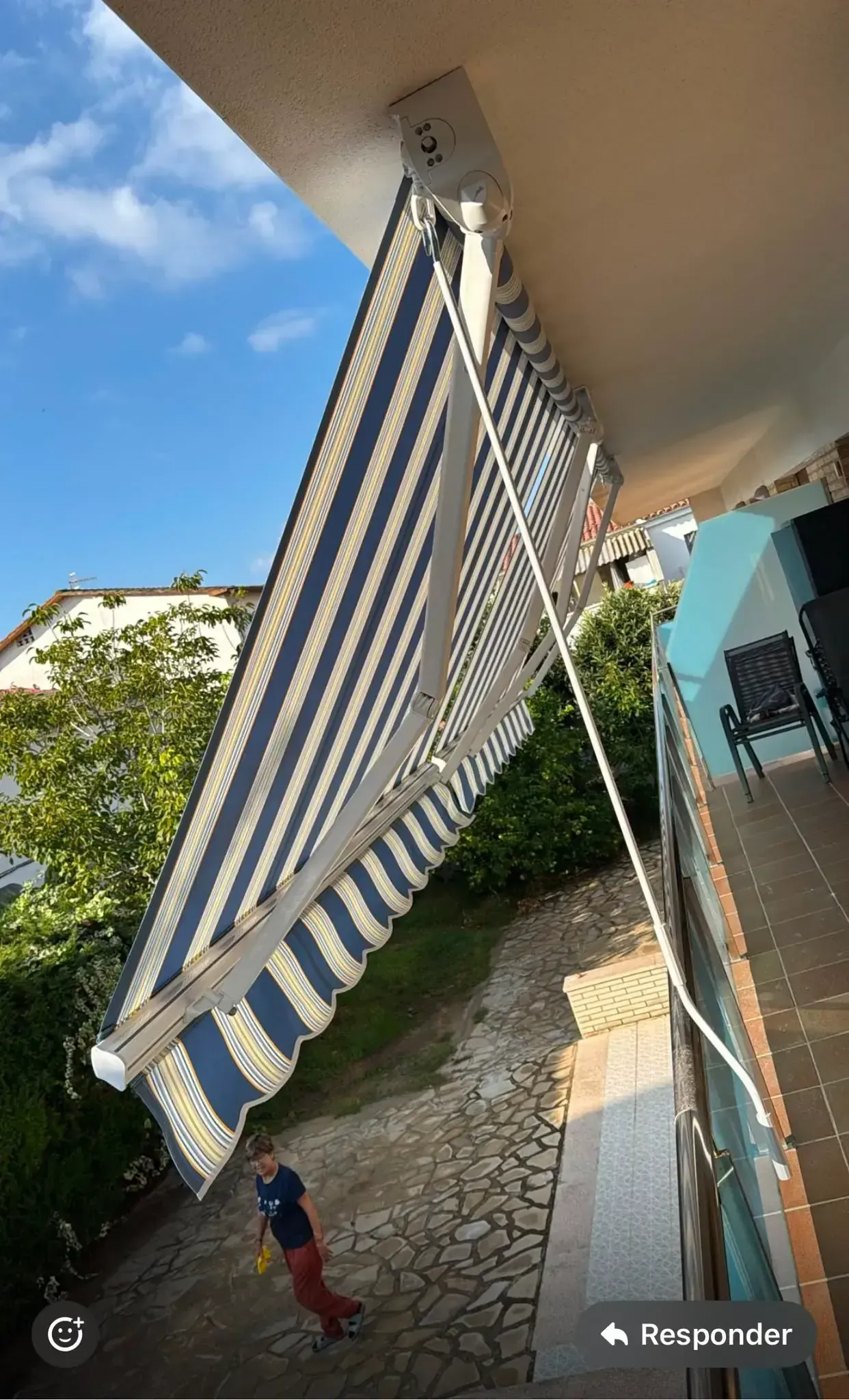 Un toldo de rayas azules y blancas se extendía sobre un patio, con un niño pequeño de pie debajo, bañado por la luz del sol.