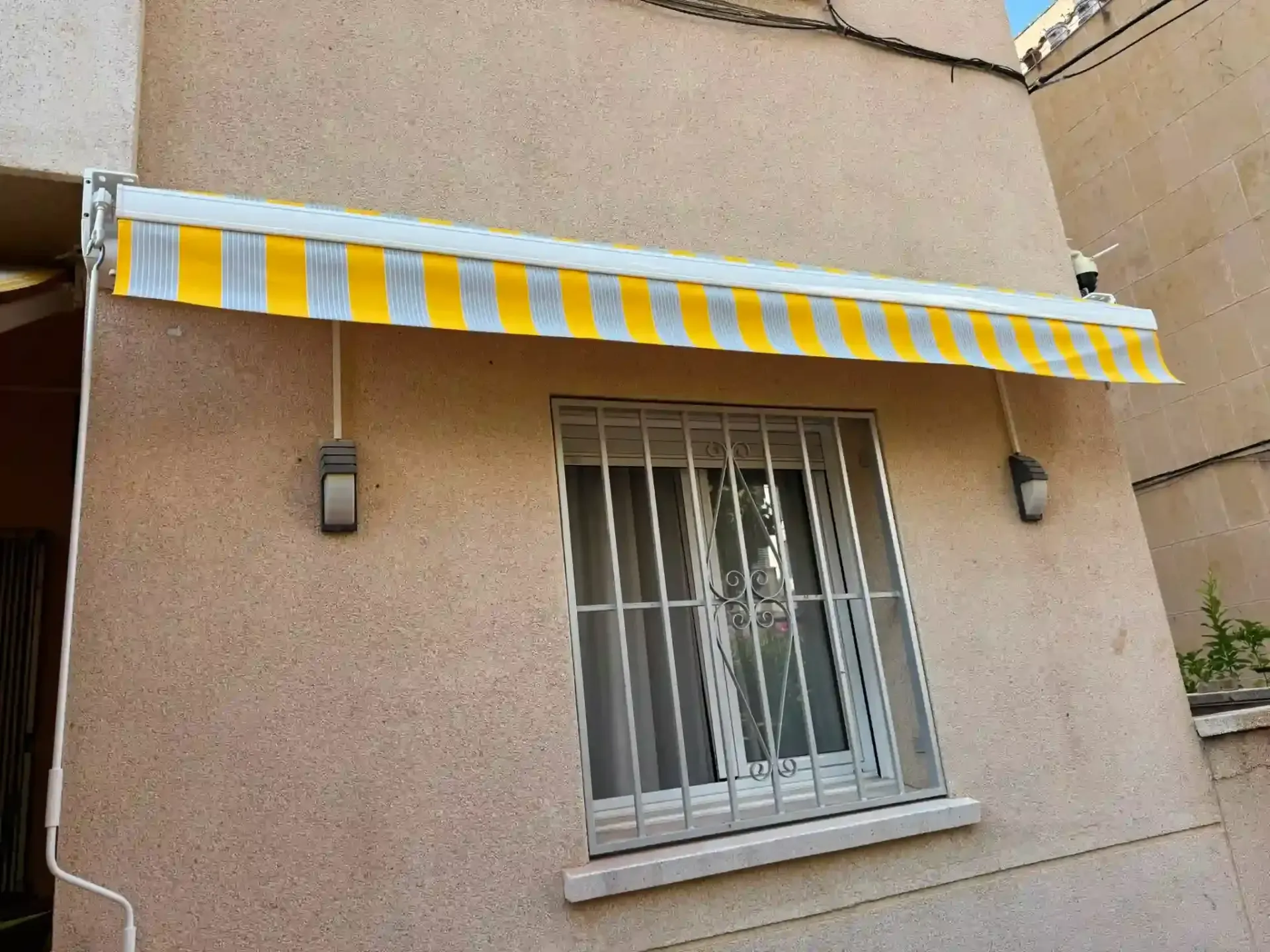 Toldo a rayas amarillas y blancas sobre una ventana enrejada en la fachada de un edificio de color beige.