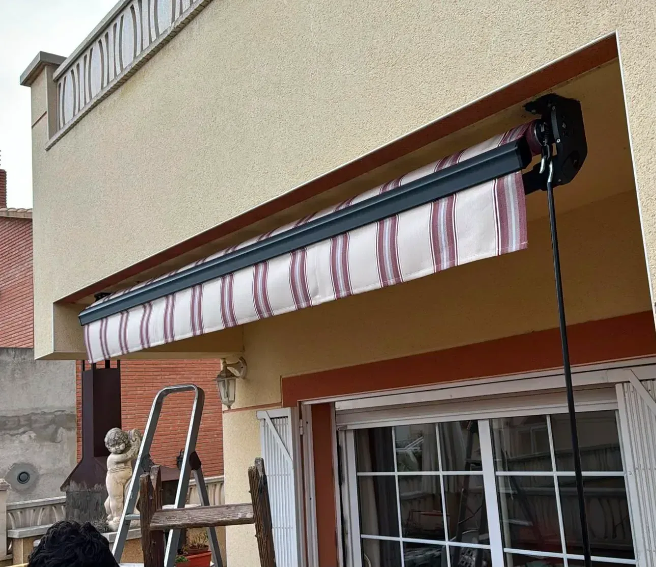 Toldo retráctil a rayas montado sobre una ventana en la fachada beige de una casa, con una escalera debajo.