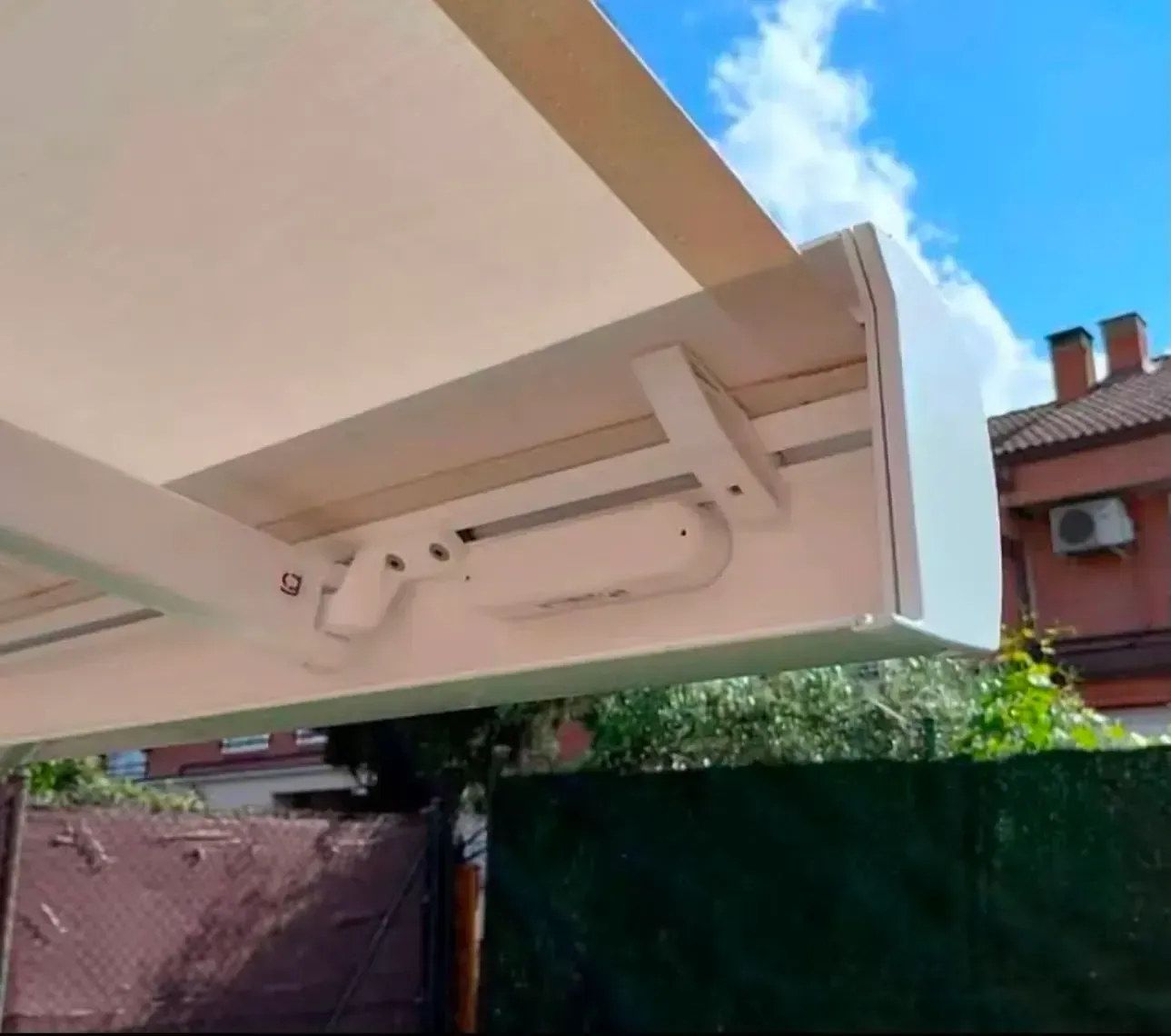 Primer plano de un soporte blanco para toldo retráctil en un patio, con árboles y tejados al fondo.