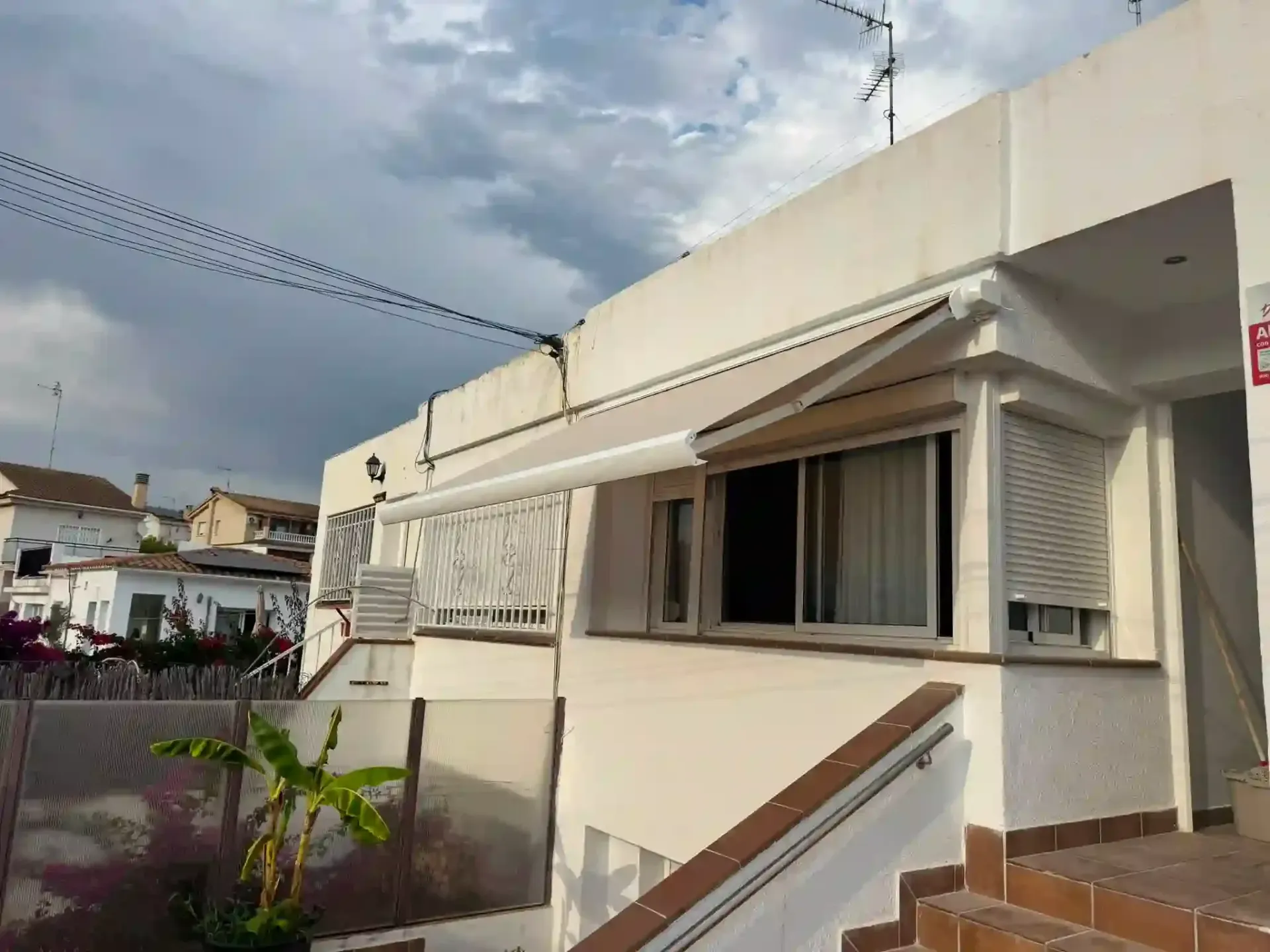 Casa de color beige con toldo y escaleras bajo un cielo nublado, vista desde una calle.