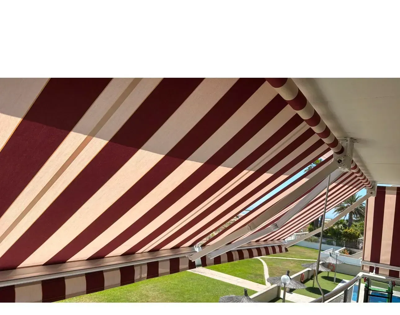 Un toldo a rayas rojas y blancas cubre un patio con césped, mesas y sillas, proyectando sombra en diagonal.