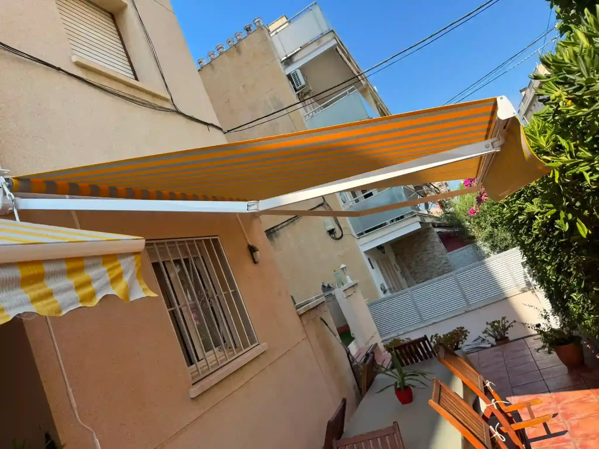 Patio con toldos a rayas naranjas, paredes de color beige y algunas macetas con plantas bañadas por la luz del sol.