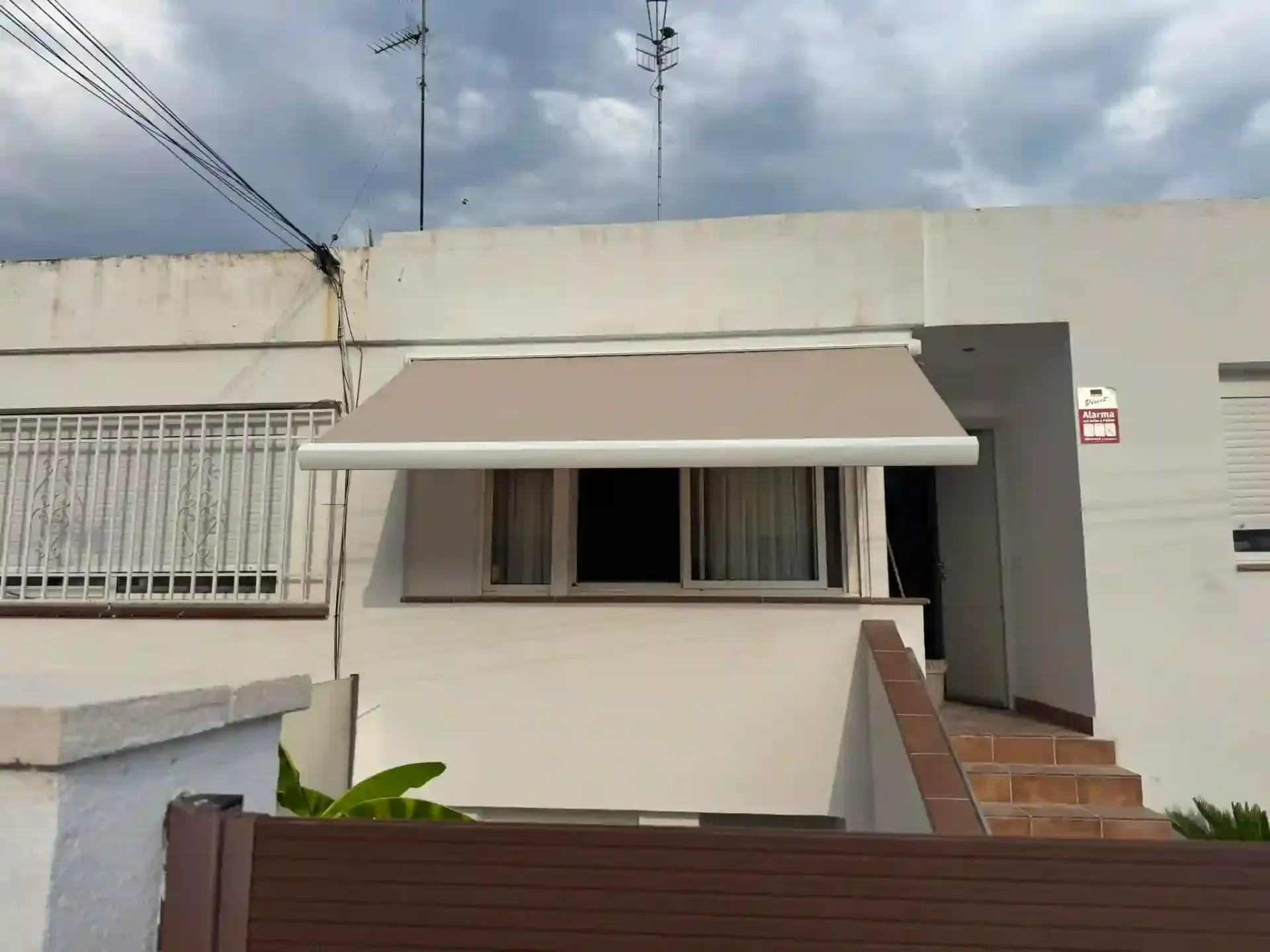 Pequeña casa blanca con toldo beige, ventana enrejada y escalones rojos bajo un cielo nublado.