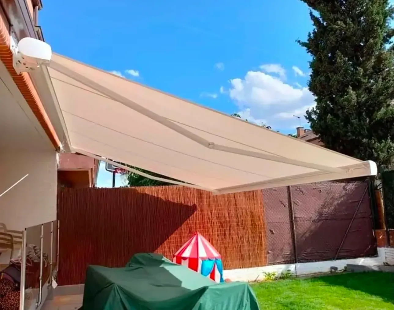 Patio trasero con toldo de vela beige, mesa cubierta con una manta verde y tienda de juegos infantil sobre el césped.