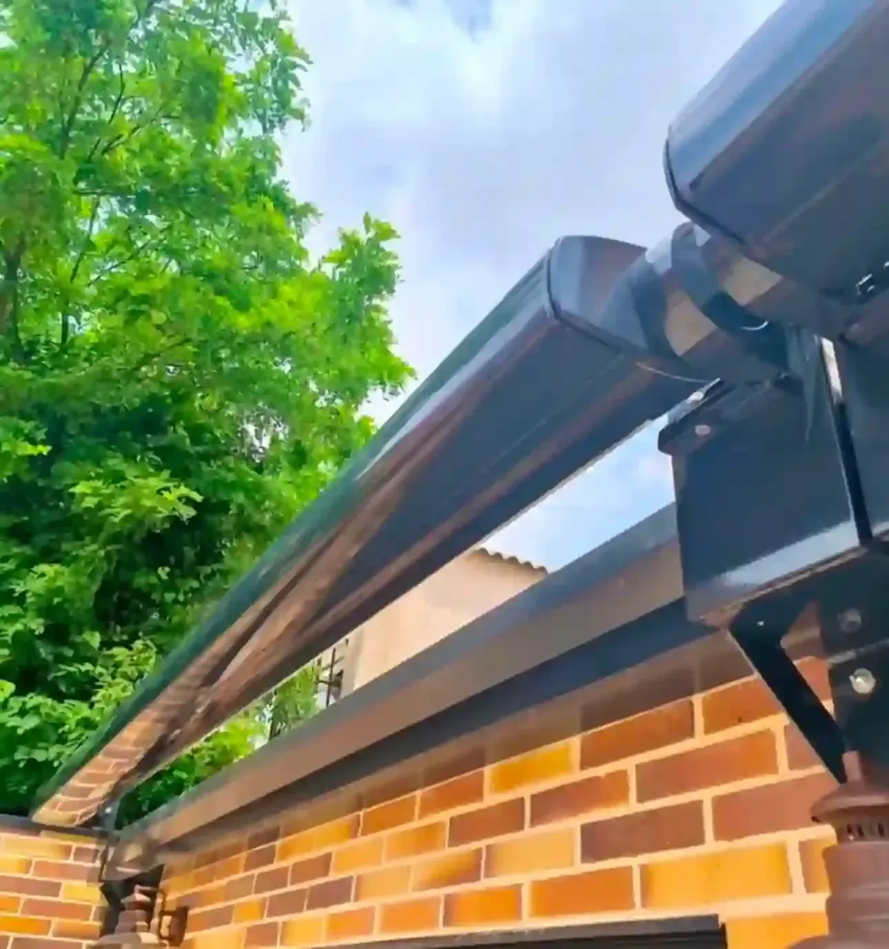 Un toldo retráctil negro se extendía sobre una pared de ladrillos bajo árboles verdes y un cielo nublado.