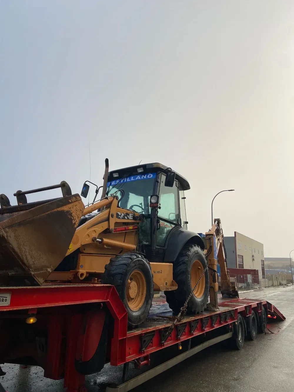 Retroexcavadora amarilla sobre un remolque rojo bajo un cielo nublado cerca de un edificio.
