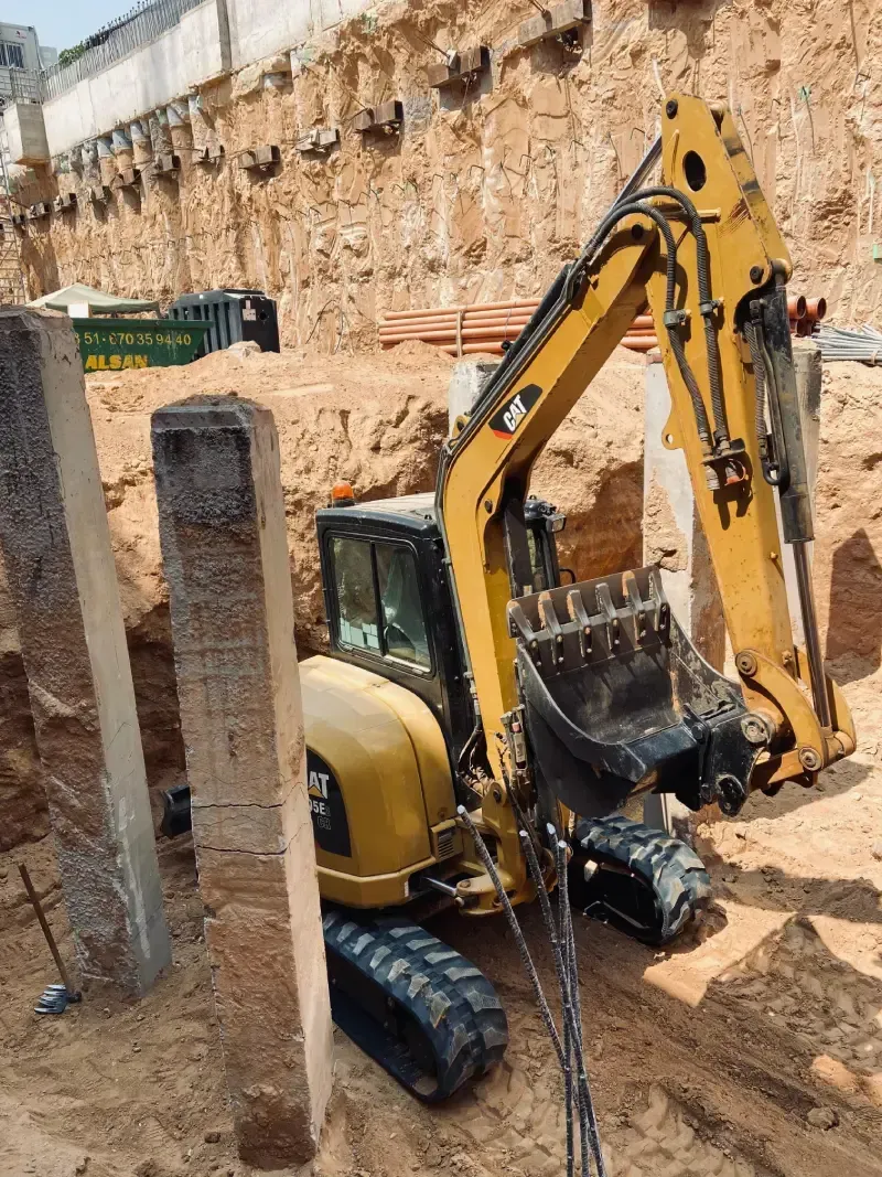 Excavadora amarilla en una excavación de un sitio de construcción, trabajando entre pilares y paredes de tierra.