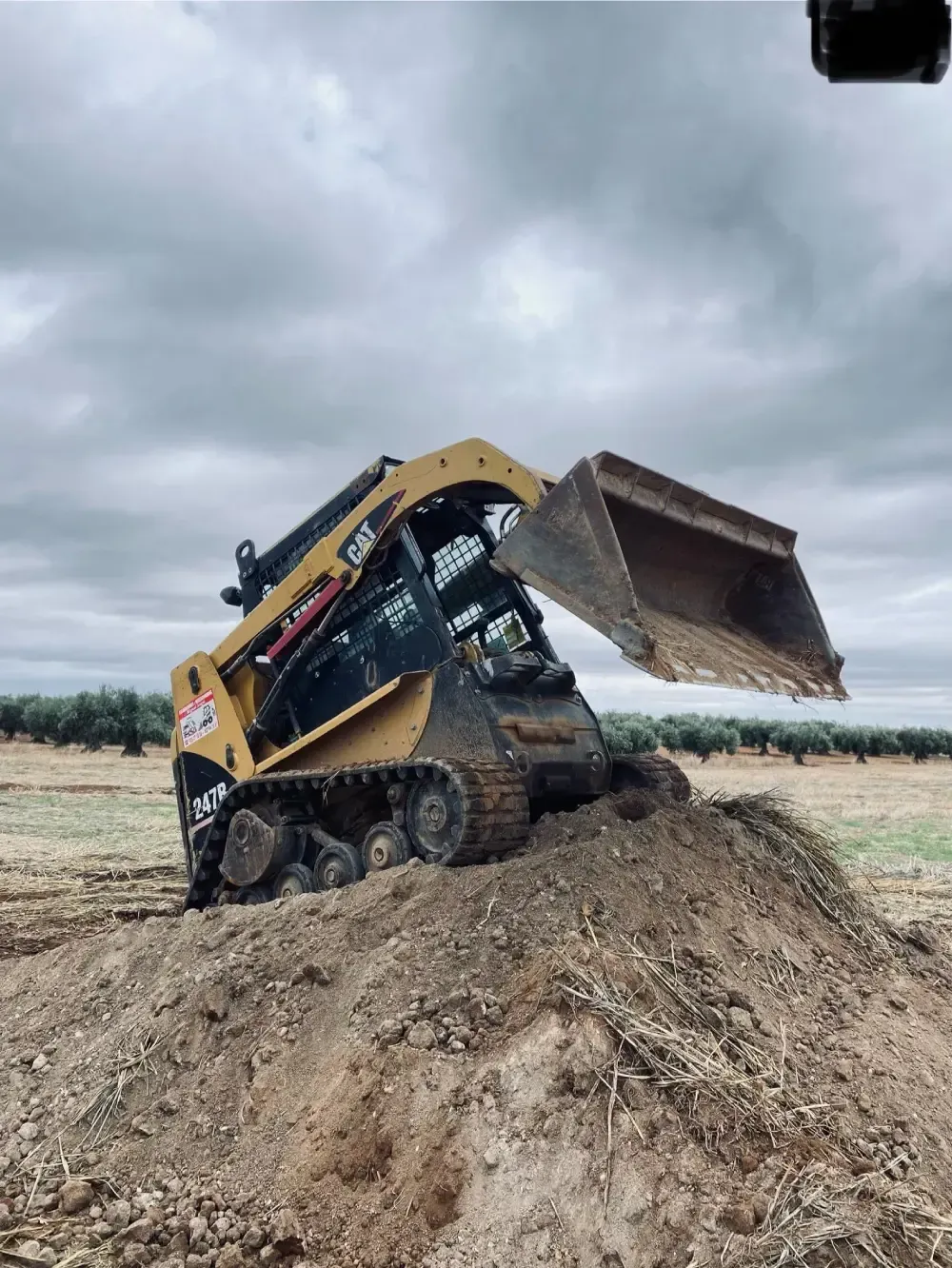 Cargadora compacta amarilla con el cucharón levantado, sobre un montón de tierra, bajo un cielo nublado.