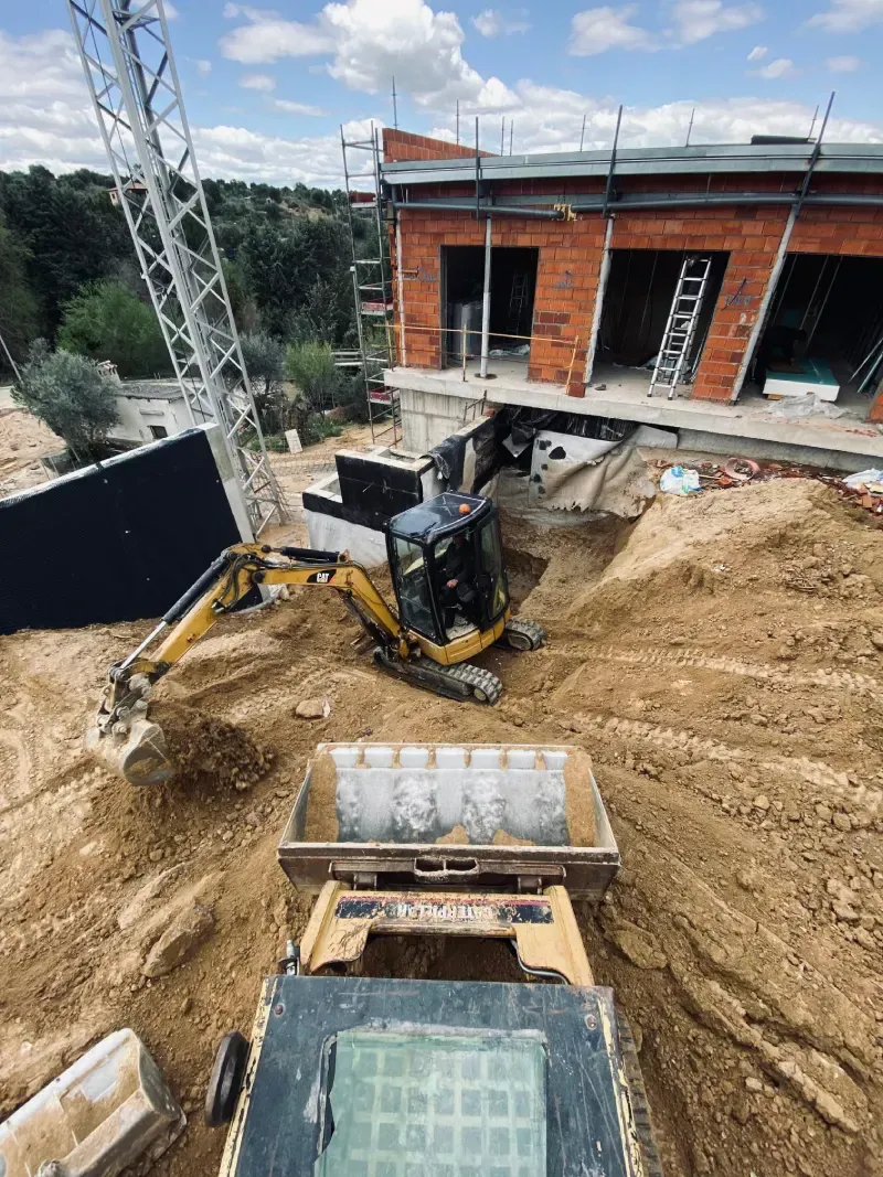 Sitio de construcción con excavadora y edificio en construcción; grúa al fondo.