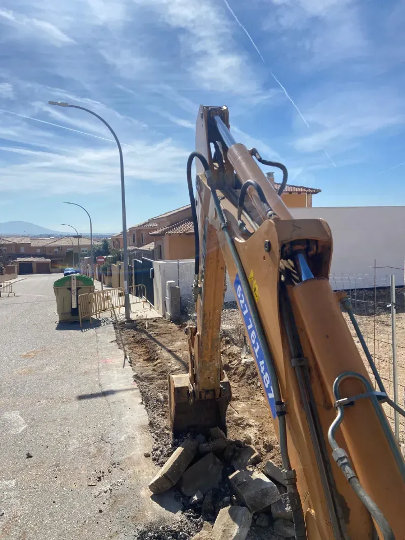 Excavadora cavando una zanja en una calle con casas al fondo bajo un cielo azul.